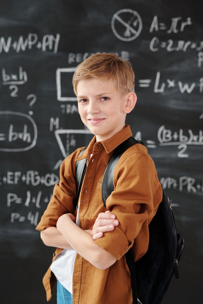 team-04 Smiling elementary student standing in classroom with math equations on blackboard.