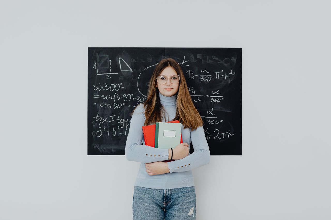 Teenage girl with glasses stands confidently in front of a blackboard with math equations, symbolizing education.